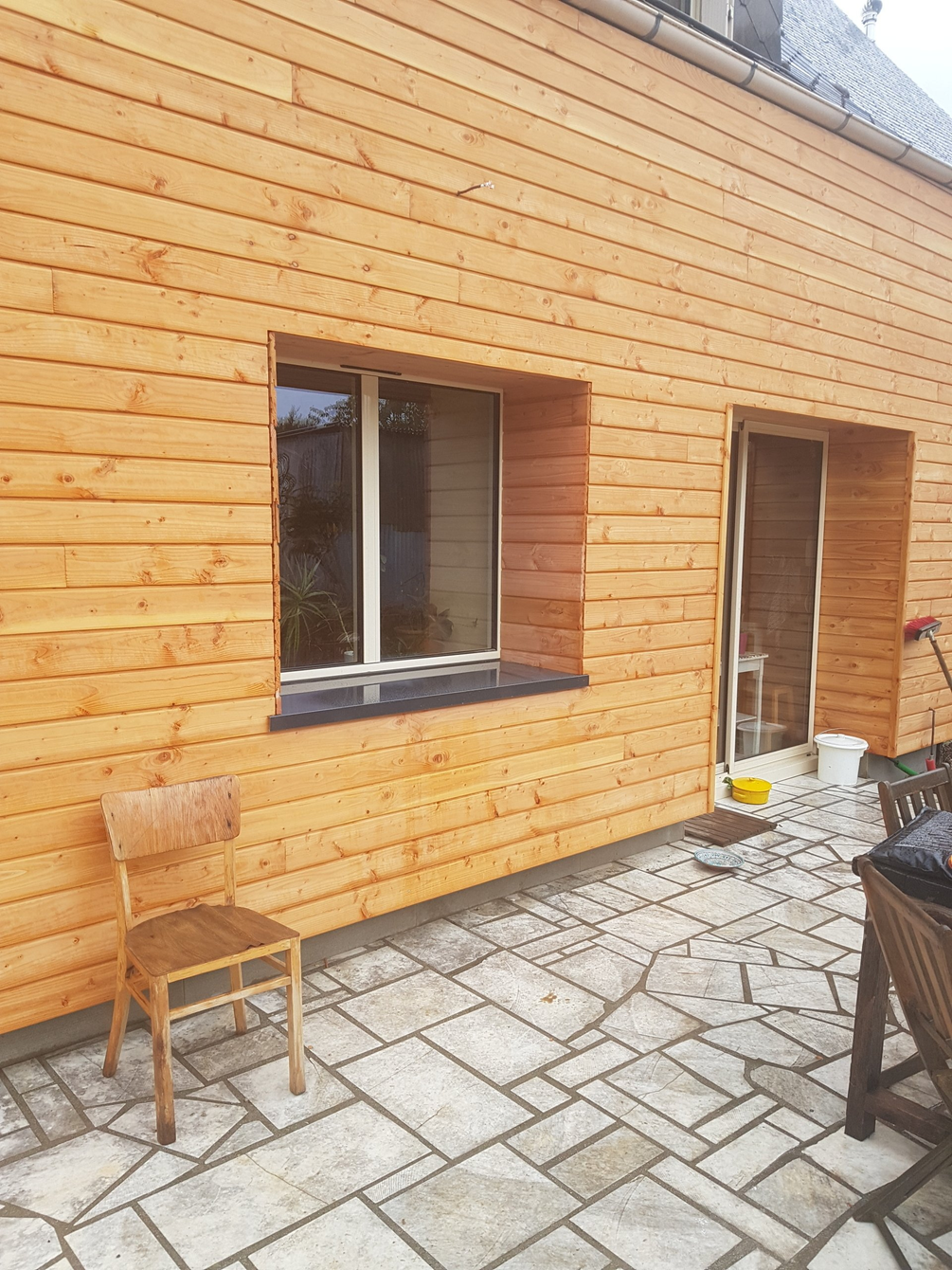 Mur en bois d'une maison avec deux fenêtres, une porte et une chaise sur une terrasse en pierre