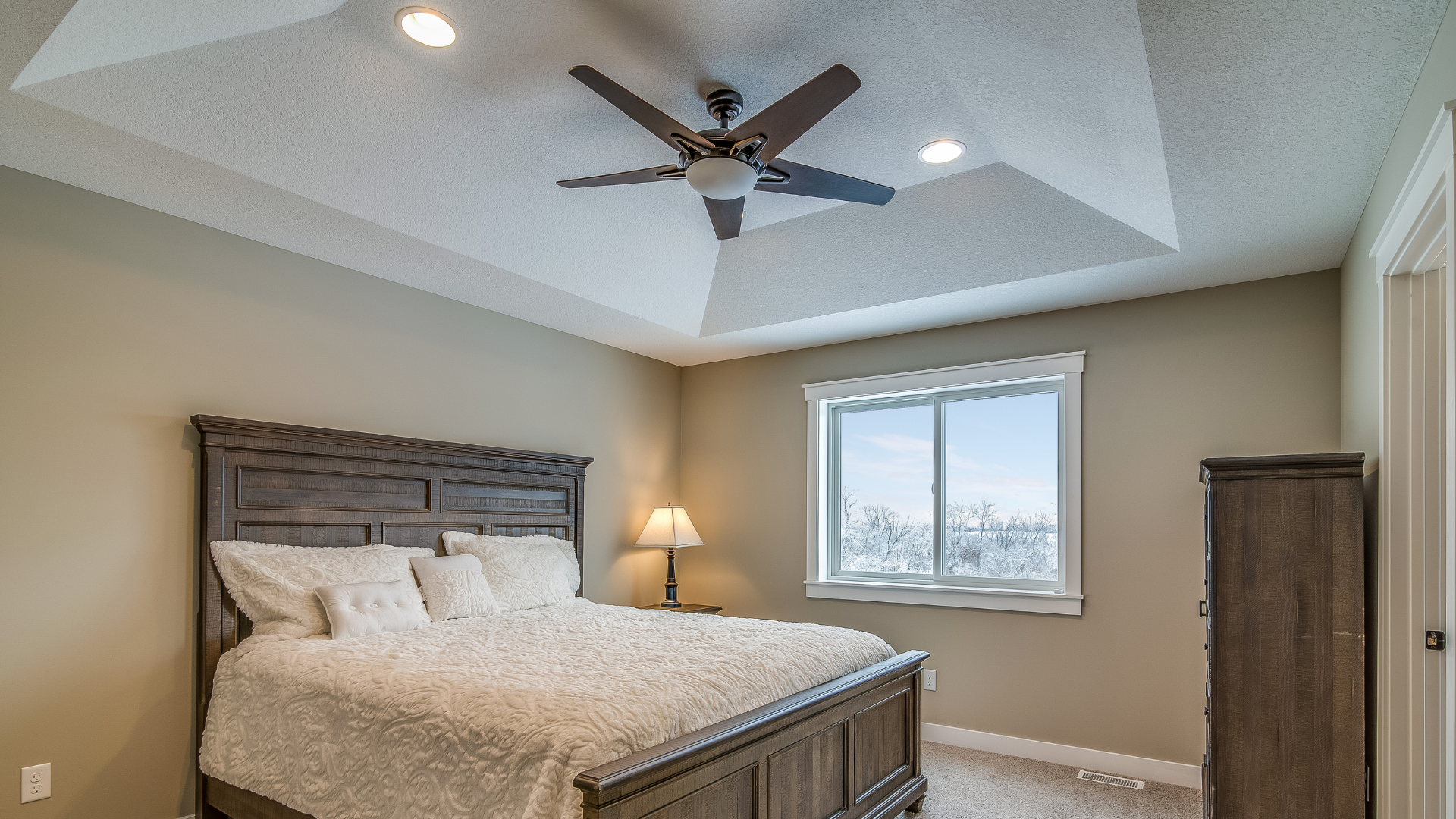 Chambre avec lit, commode, fenêtre et ventilateur de plafond, murs et moquette beiges.