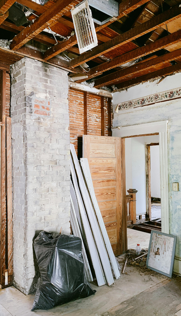 Pièce intérieure en rénovation, poutres apparentes, cheminée en briques, débris, portes et un sac-poubelle noir.
