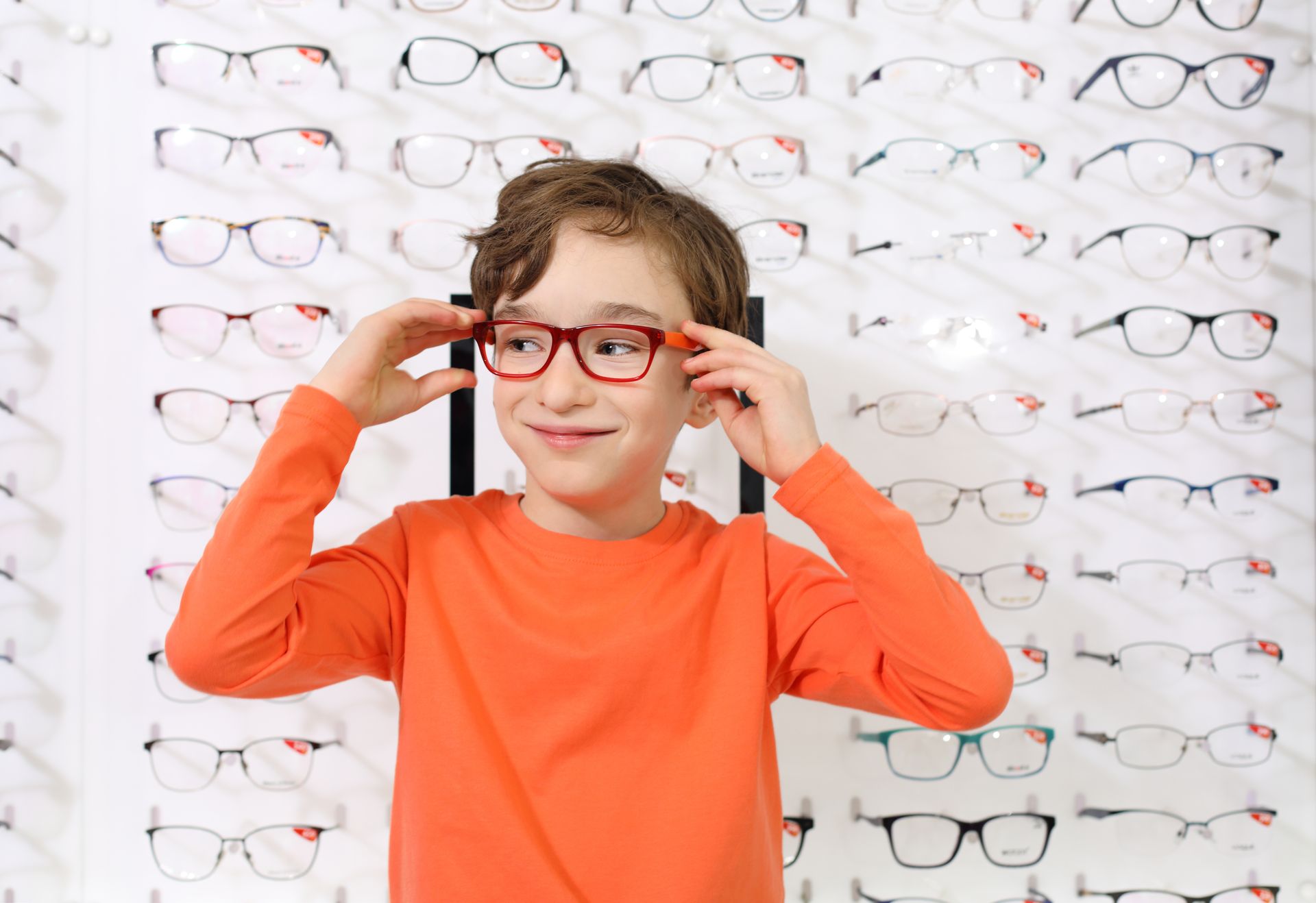 Garçon en chemise orange essayant des lunettes rouges dans un magasin d'optique, entouré de montures.