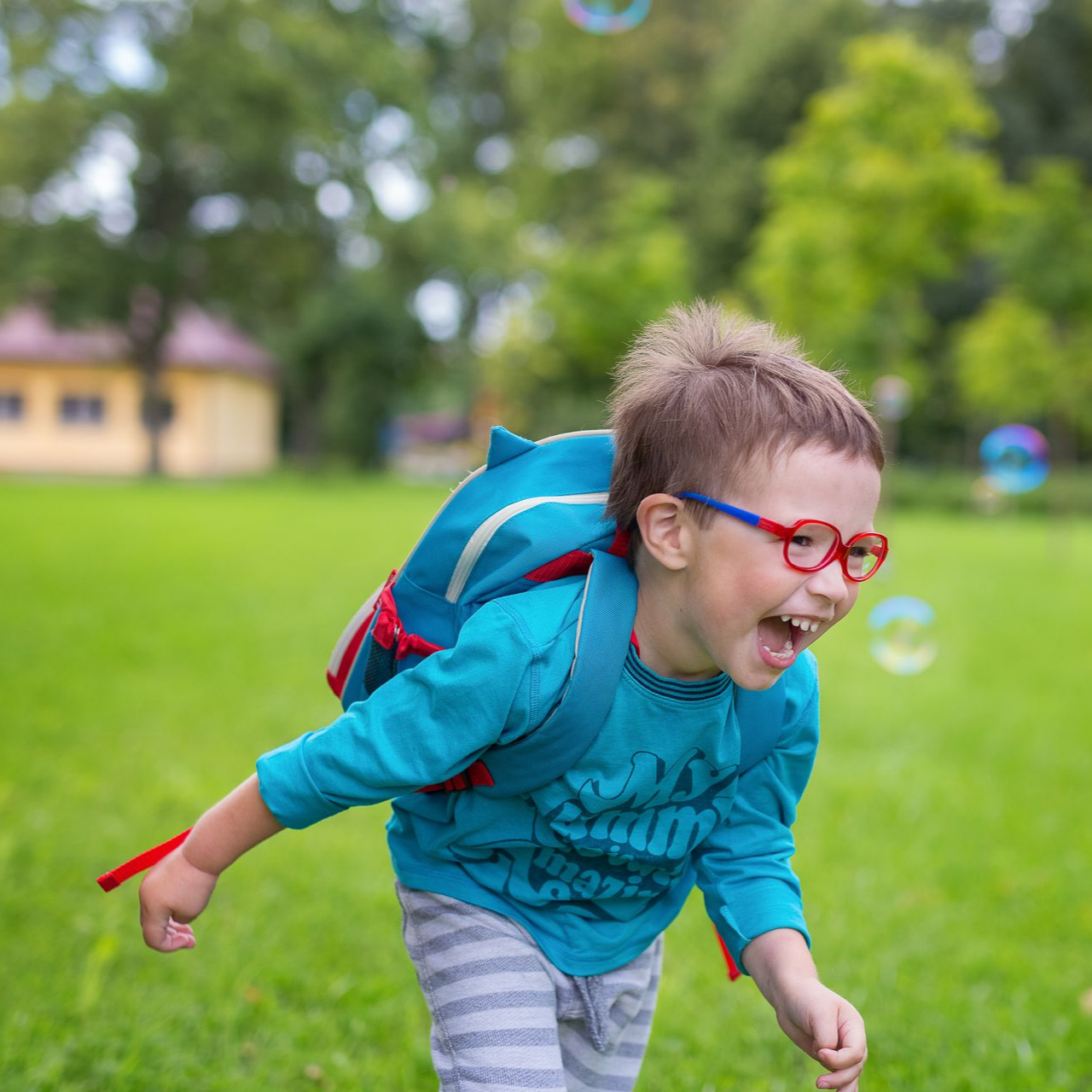 Un garçon portant des lunettes rouges rit dehors, sur une pelouse verdoyante où il y a des bulles.