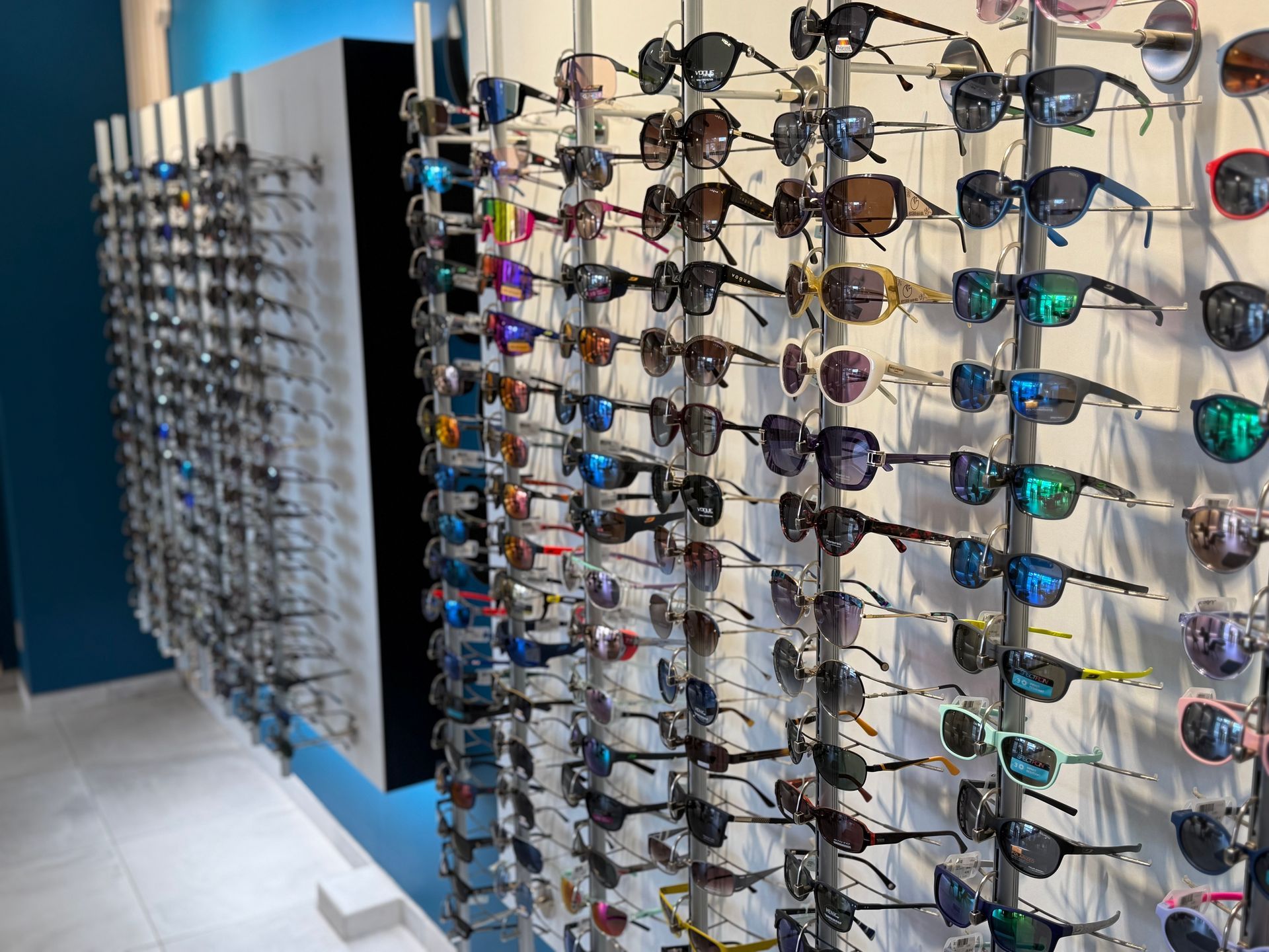 Des lunettes de soleil de styles et de couleurs variés sont exposées sur un mur blanc dans un magasin.