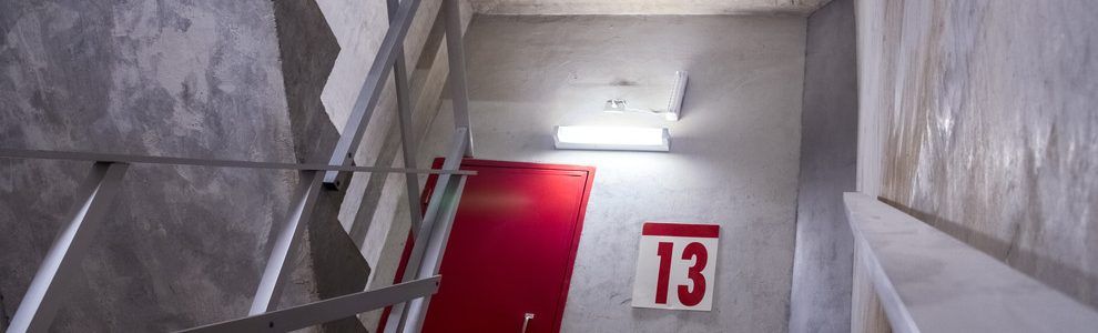 Cage d'escalier avec porte rouge et numéro 13. Murs en béton et rampes métalliques. Éclairage fluorescent au plafond.