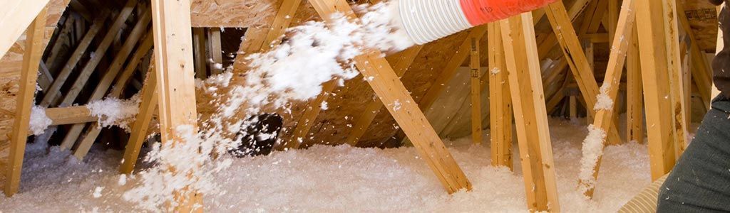 Une personne gantée applique de l'isolant en vrac dans un grenier. On aperçoit la charpente en bois et l'isolant blanc.