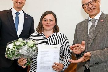 Zwei Männer und eine Frau stehen nebeneinander und halten Blumen und ein Stück Papier in den Händen.