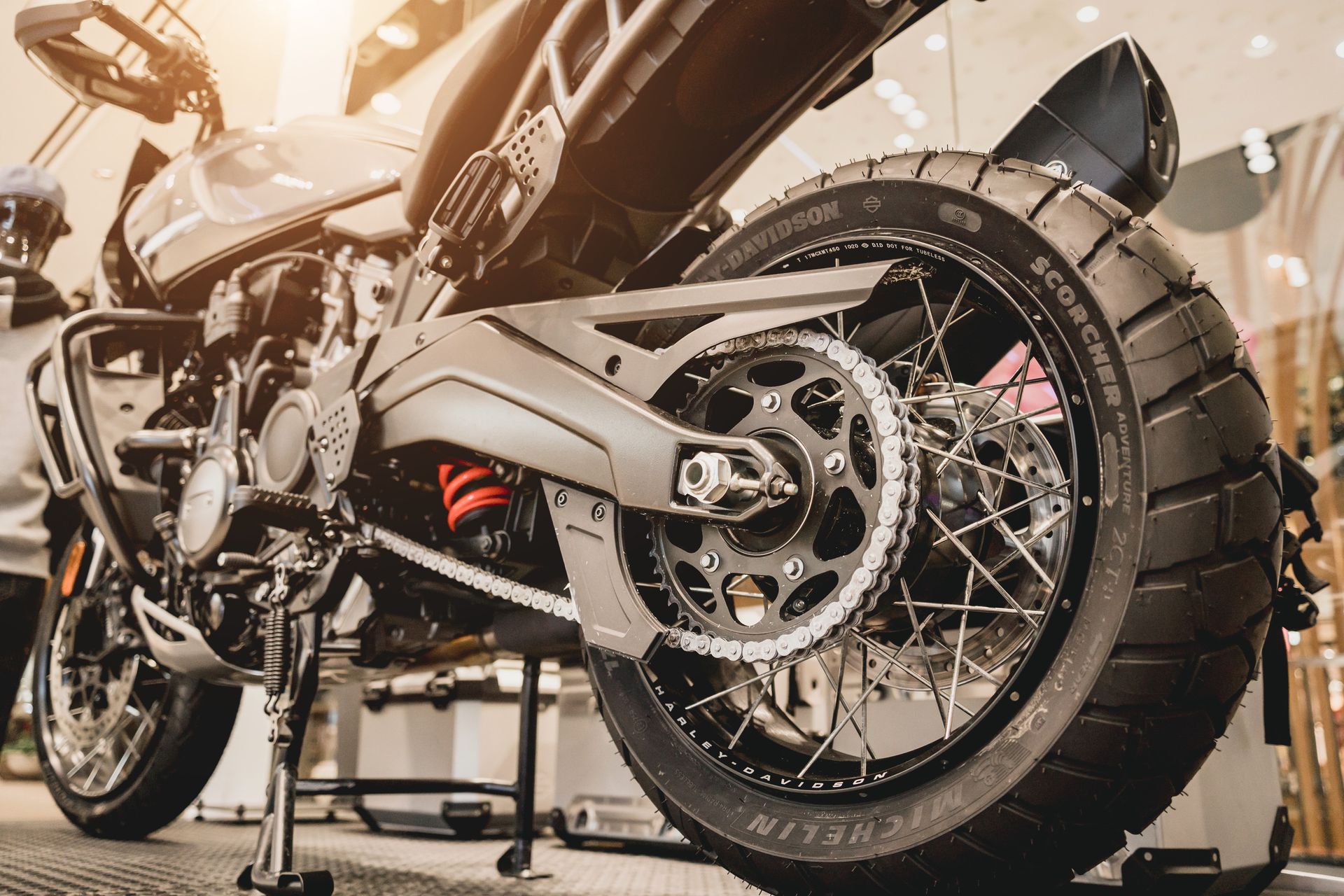 Primer plano de la parte trasera de una motocicleta gris con un neumático negro y una cadena contra un fondo interior borroso.