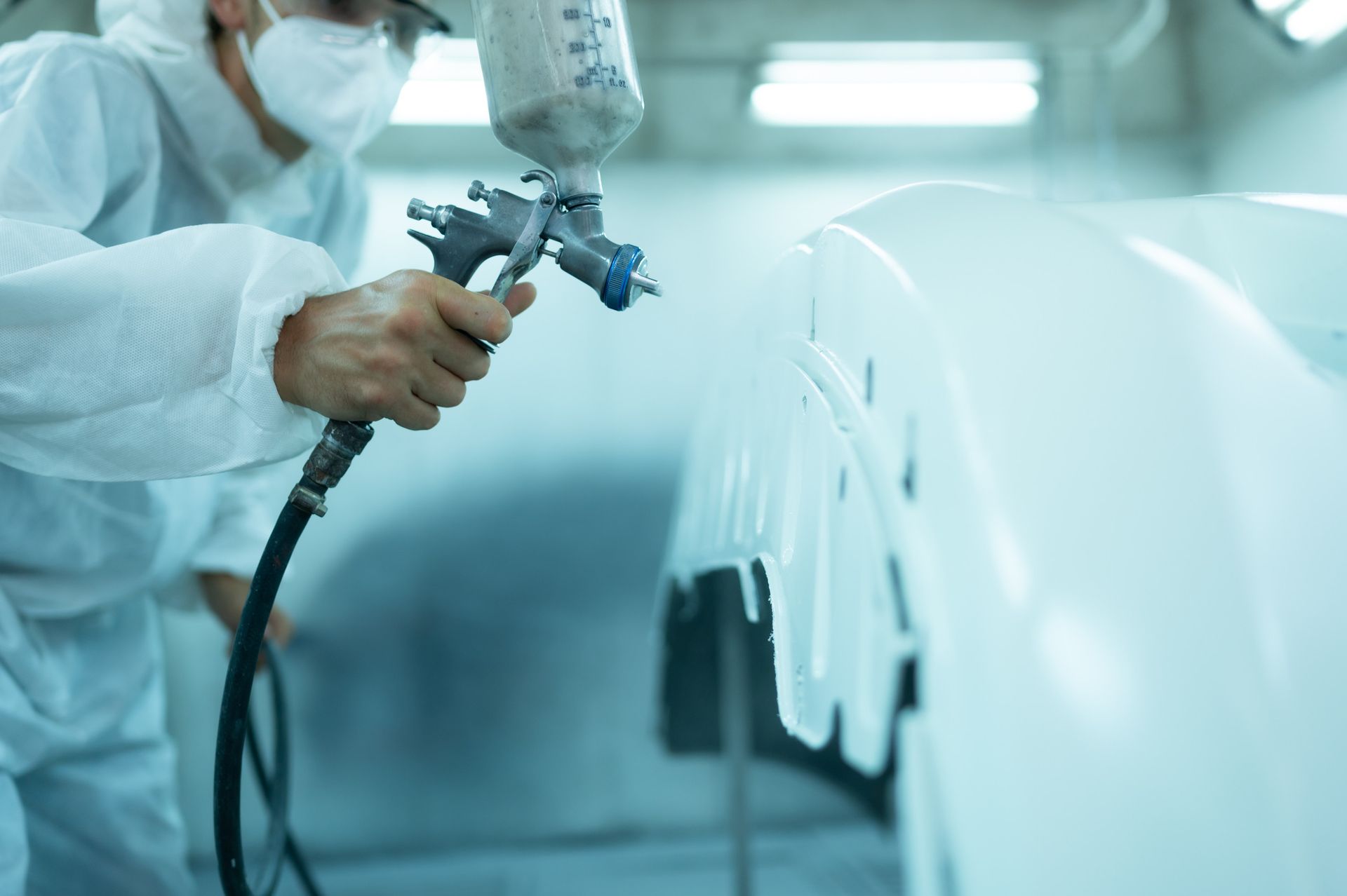 Persona con traje de protección pulveriza pintura blanca sobre el parachoques de un coche en un taller.