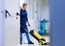Una mujer con uniforme azul pasa la aspiradora por un pasillo; se ve un carrito de limpieza.