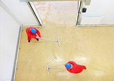 Dos personas con trajes rojos y sombreros azules secan con una escobilla de goma un suelo mojado en el interior de una puerta.