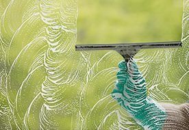 Mano con guante verde usando una espátula sobre una ventana jabonosa, limpiando con fondo verde.