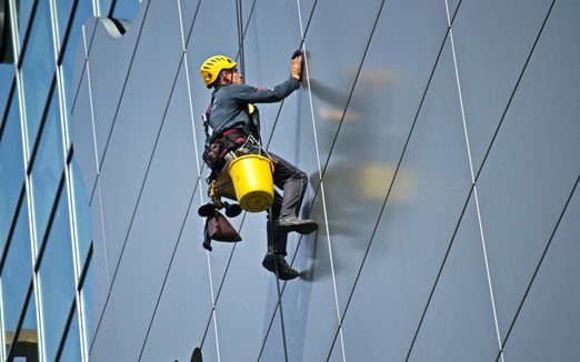 Limpiador de ventanas en un edificio de gran altura; sujeto con arnés, lavando un panel de vidrio.