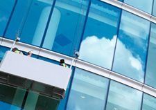 Limpiadores de ventanas en una plataforma blanca limpiando un rascacielos de cristal reflectante; cielo azul con nubes.