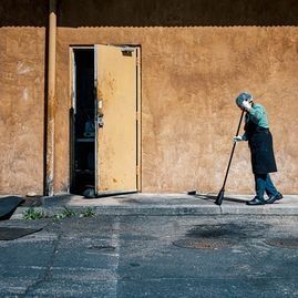 Una persona mayor con delantal barre la acera cerca de una puerta amarilla contra una pared naranja texturizada.
