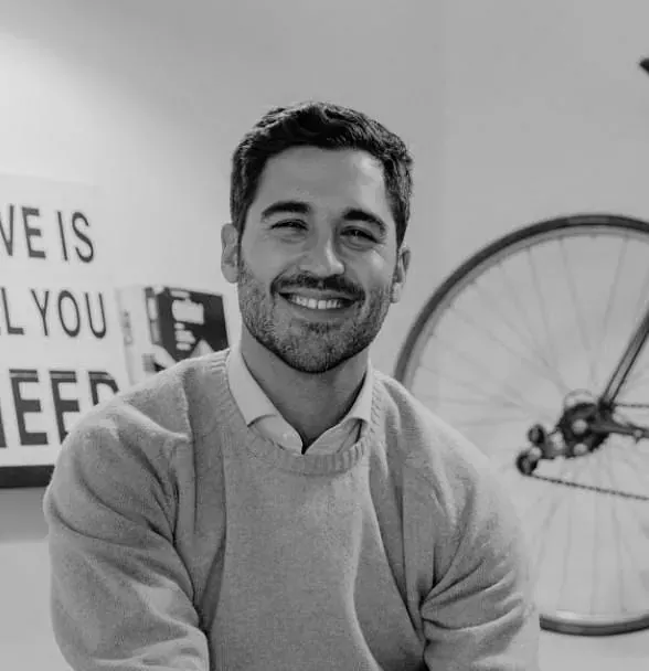 Hombre sonriente vestido con un suéter de color claro, sentado cerca de una rueda de bicicleta y un cartel en la pared que dice 