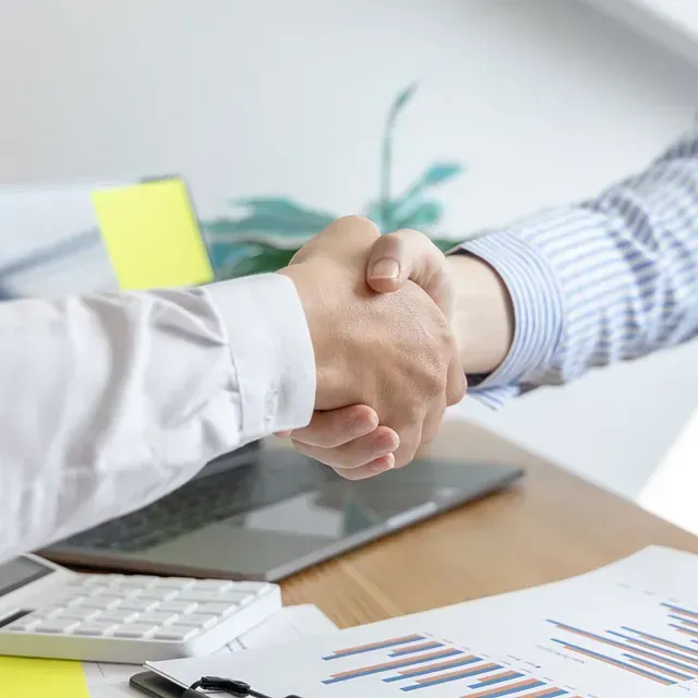 Dos personas vestidas de negocios se dan la mano sobre un escritorio con papeles y una computadora portátil.