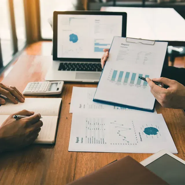 Dos personas revisan datos financieros en una mesa, utilizando una computadora portátil, gráficos, una calculadora y un cuaderno.
