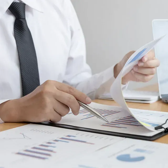 Persona con camisa blanca y corbata analizando informes financieros con un bolígrafo en un escritorio con una computadora portátil.