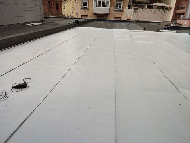 Material de techado blanco siendo instalado sobre un tejado plano, con un fondo de edificios urbanos.