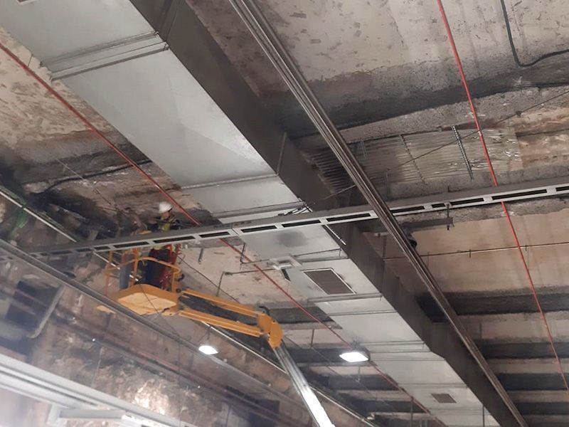 Trabajador en un ascensor instalando conductos eléctricos en un techo con estructura metálica. Entorno industrial.