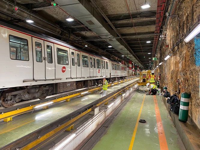 Tren subterráneo en un túnel de mantenimiento, con trabajadores cerca. Las paredes del túnel son ásperas, rocosas y poco iluminadas.