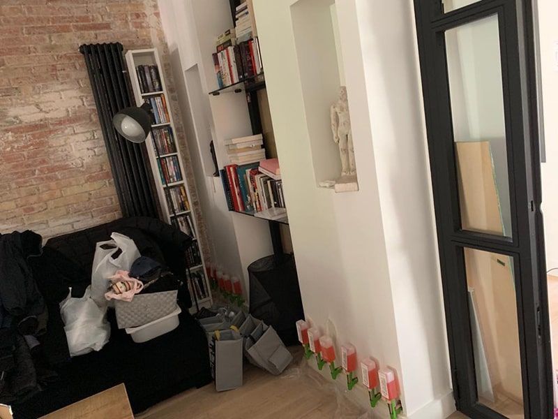 Sala de estar desordenada con un sofá negro, una estantería y una puerta con marco negro.