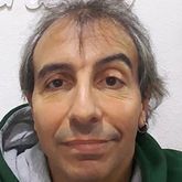 Hombre de cabello canoso, expresión neutra, vestido con una sudadera con capucha verde y un pendiente.