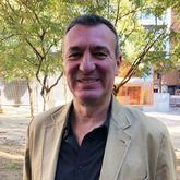 Hombre con cabello castaño corto, sonriente, vestido con una chaqueta color canela y una camisa negra al aire libre cerca de árboles y un edificio.