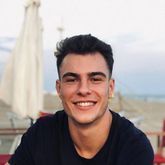 Un joven de cabello oscuro, con una amplia sonrisa. Lleva una camisa negra y está sentado al aire libre con la playa de fondo.