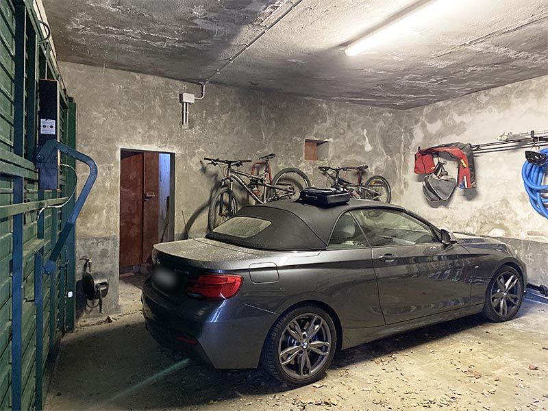 Un automóvil convertible gris oscuro estacionado en un garaje desordenado con bicicletas, herramientas y una puerta abierta.