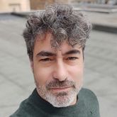 Hombre con cabello gris rizado y barba, mirando a cámara, con un suéter verde, en una azotea.