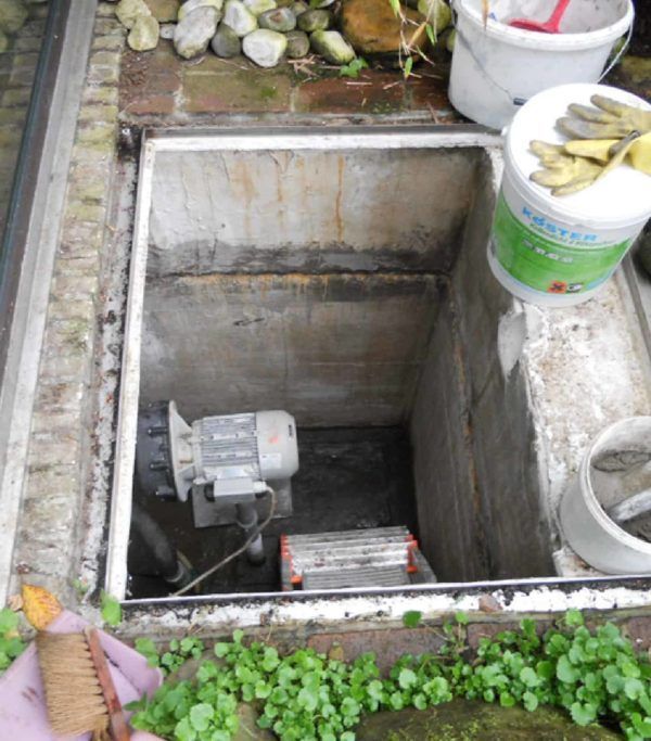 Pozo de hormigón abierto con bomba, escalera pequeña y herramientas; el entorno incluye vegetación, un balde y guantes.