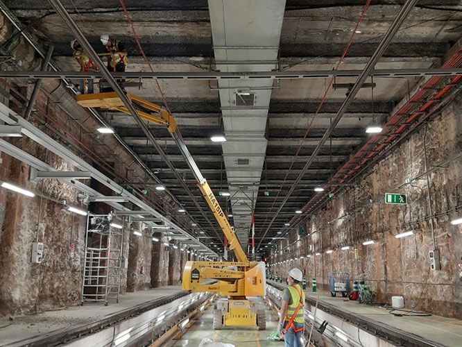 Trabajadores en un túnel con maquinaria, instalando infraestructura. Ascensor amarillo, paredes de roca expuestas, iluminación cenital.
