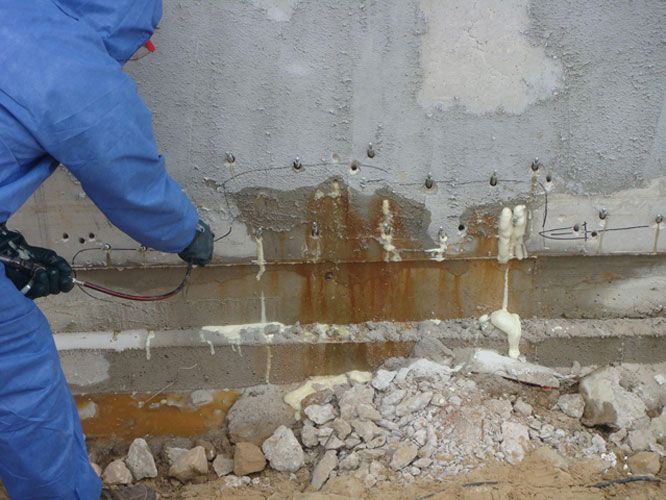 Una persona con traje azul de protección rocía sellador de espuma sobre una pared de hormigón manchada. Agua marrón corre por la pared.