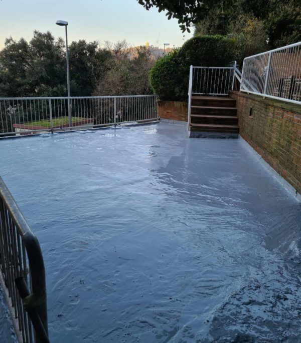 Patio exterior pintado de gris con barandillas y escaleras de metal, una pared de ladrillo y árboles al fondo.