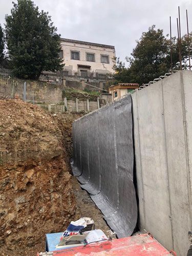 Obra con muro de hormigón, membrana impermeabilizante negra y tierra excavada. Edificio al fondo.