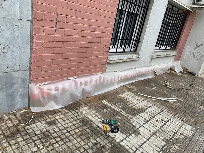 Exterior del edificio con pared de ladrillo rosa, ventana con rejas, lona de plástico y herramientas sobre suelo de baldosas.