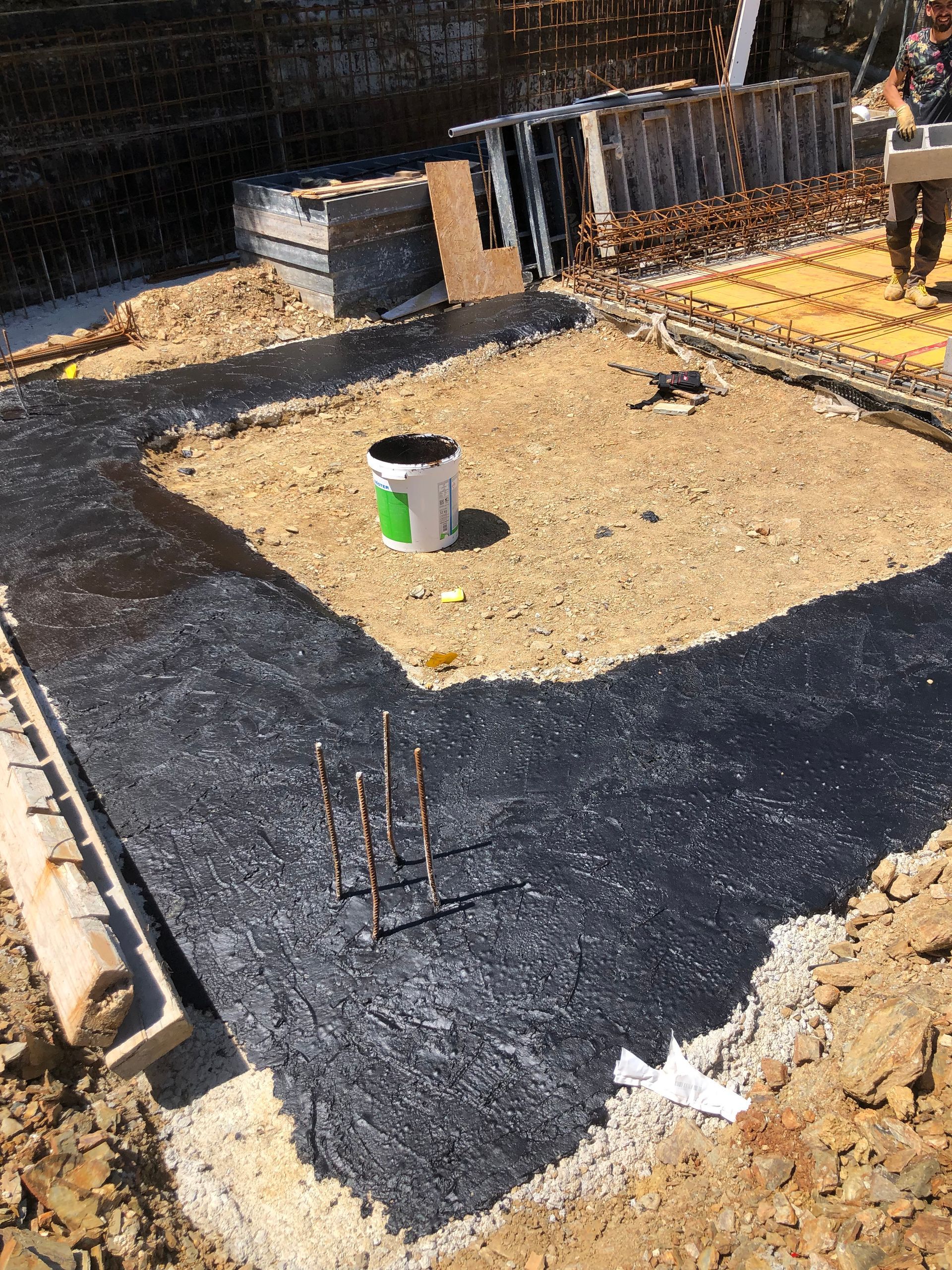 Sitio de construcción con base de cimentación oscura, balde, varilla de refuerzo y persona caminando.