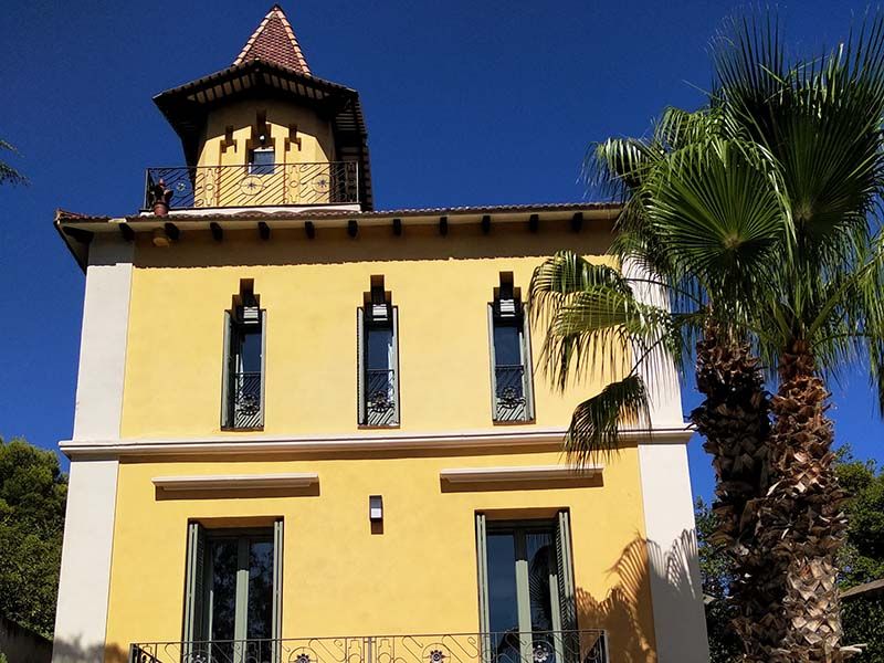 Edificio amarillo con torre, ventanas arqueadas, palmera y cielo azul.