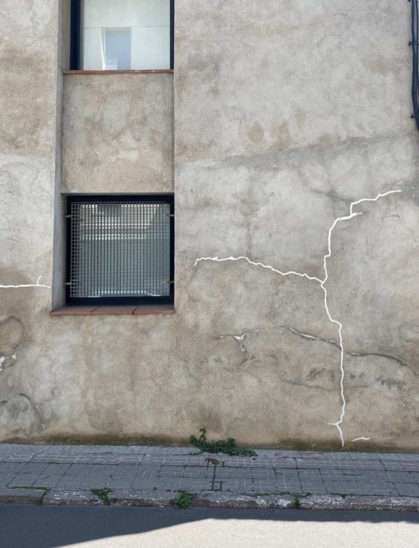 Fachada de edificio gris con dos ventanas, una de ellas con reja de seguridad; gran grieta recorre la pared.
