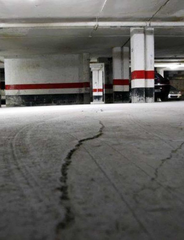 Piso de hormigón agrietado en un estacionamiento subterráneo, con columnas de soporte marcadas con rayas rojas y negras.