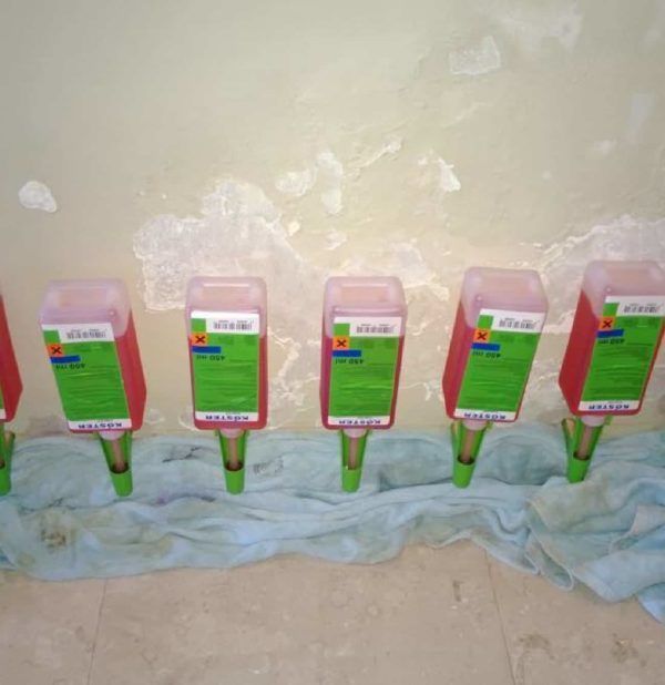 Seis botellas rojas de pesticidas montadas en una pared, apoyadas sobre caños verdes y sobre una tela azul.