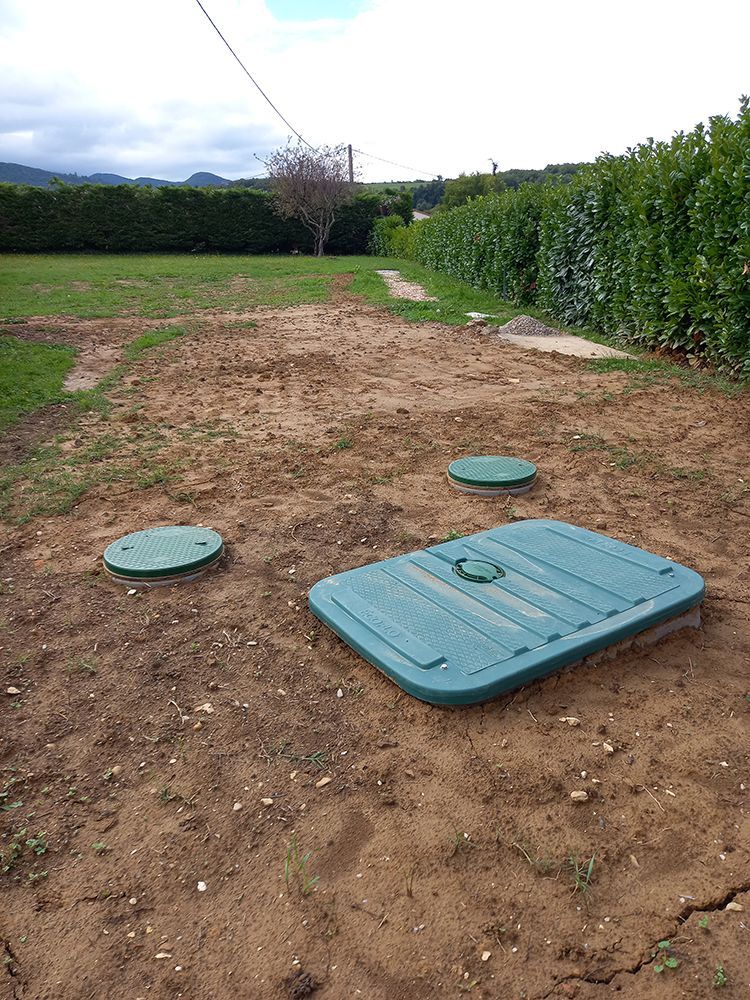 Trois couvercles de fosse septique verts sur une cour en terre, avec une zone herbeuse et des haies en arrière-plan.