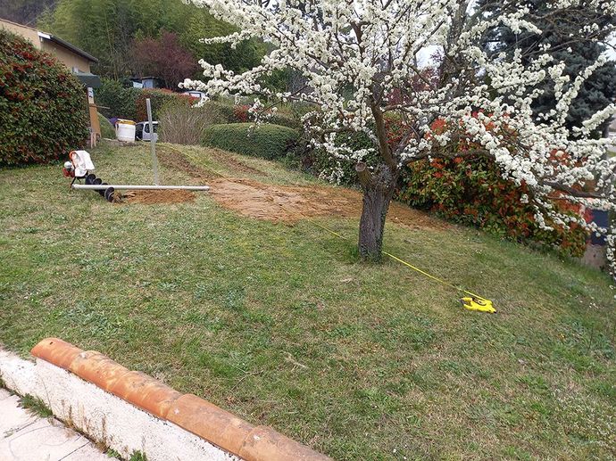 Pelouse verte avec un arbre en fleurs; une petite tranchée creusée, des machines et une corde jaune présentes.