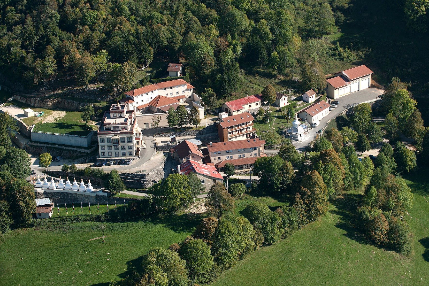 Vue aérienne d'un village niché dans une vallée verdoyante et arborée, avec des bâtiments, des routes et un champ herbeux.