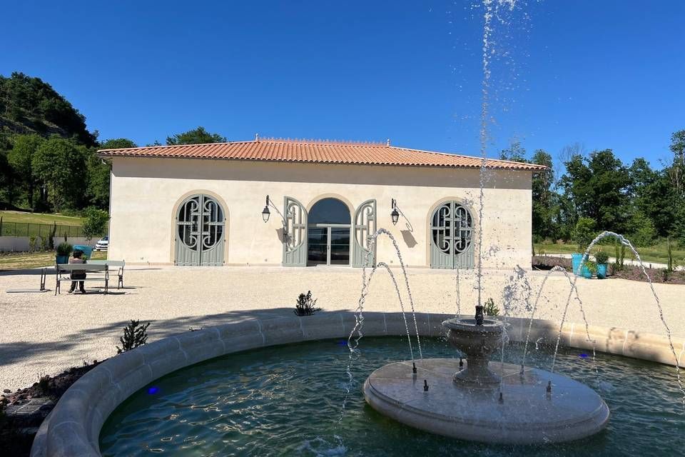 Un bâtiment en pierre avec des fenêtres cintrées et un toit en terre cuite se trouve près d'une fontaine sous un ciel bleu.
