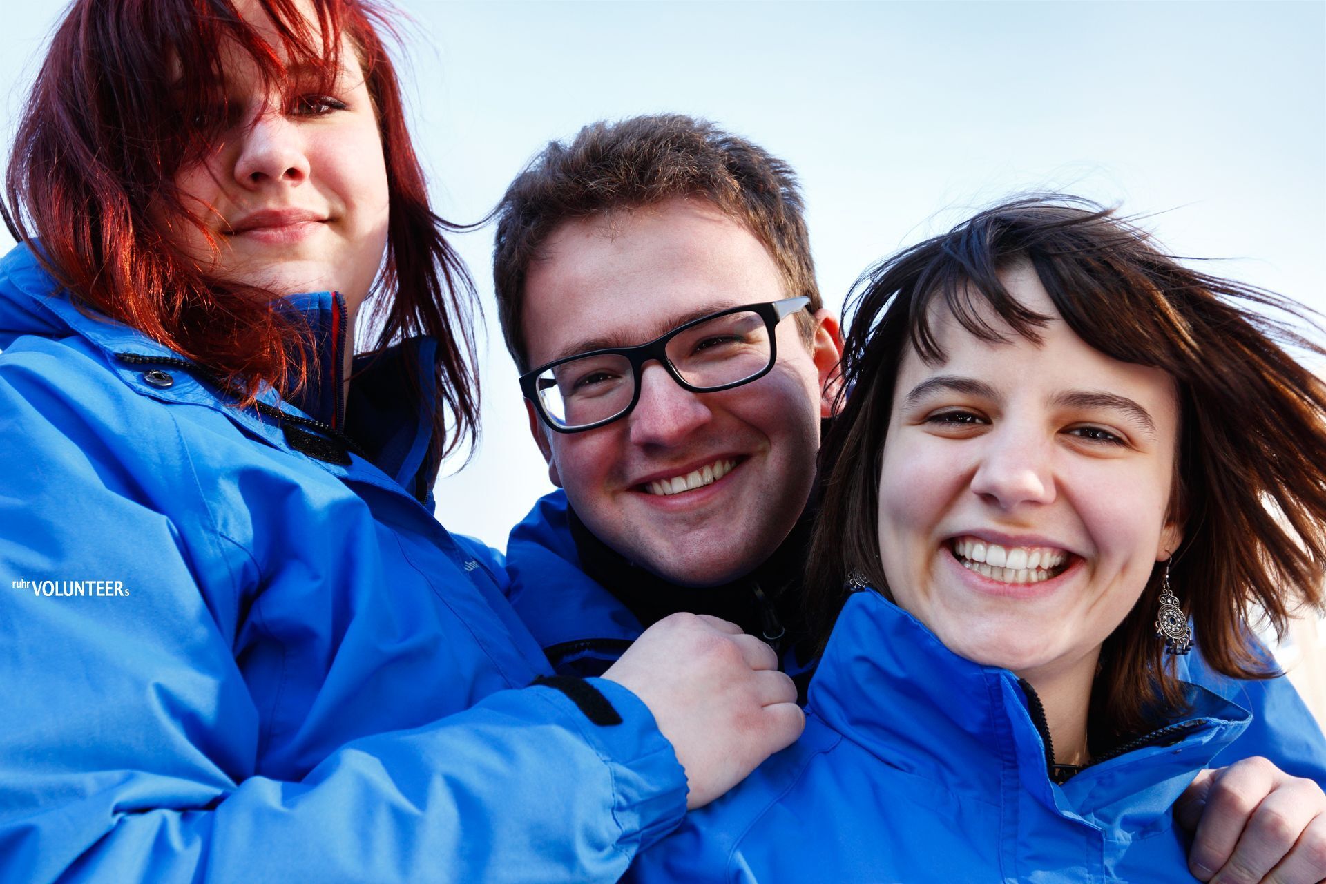 Ein Mann und zwei Frauen in blauen Jacken lächeln in die Kamera