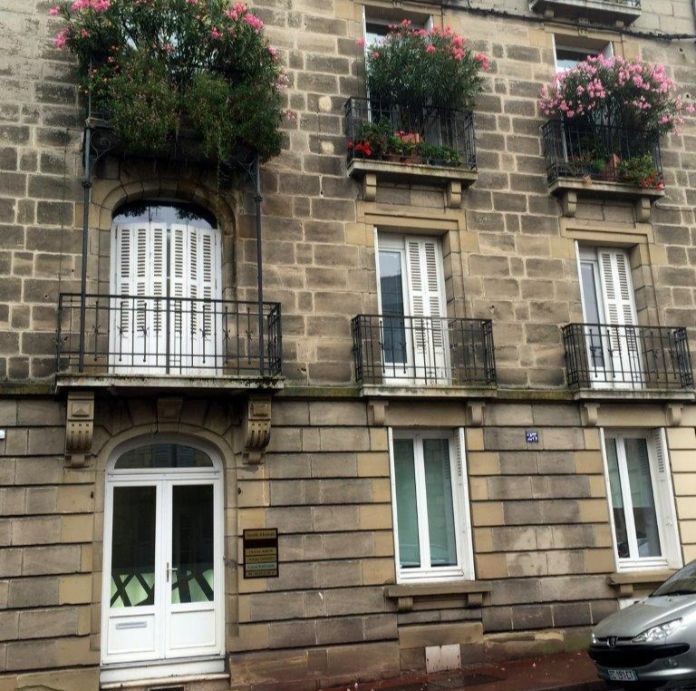 Immeuble en pierre avec balcons, fenêtres à volets et jardinières ; une voiture garée est visible.