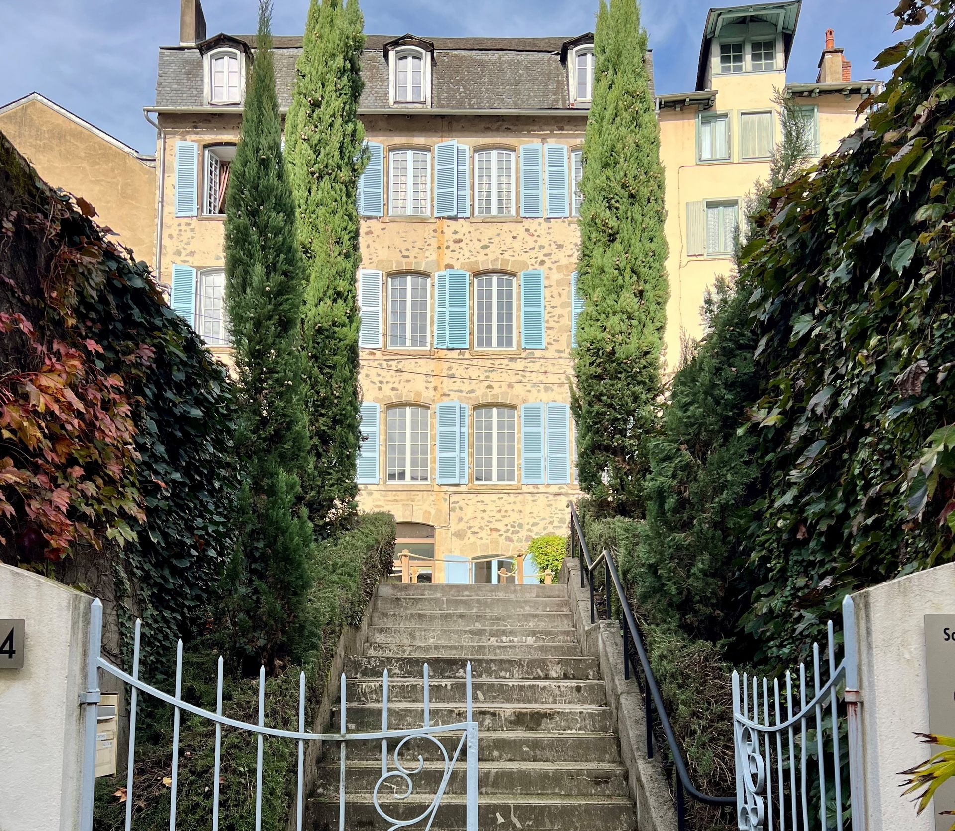 Bâtiment en pierre avec des volets bleu clair, de grands arbres et un escalier menant à l'étage.