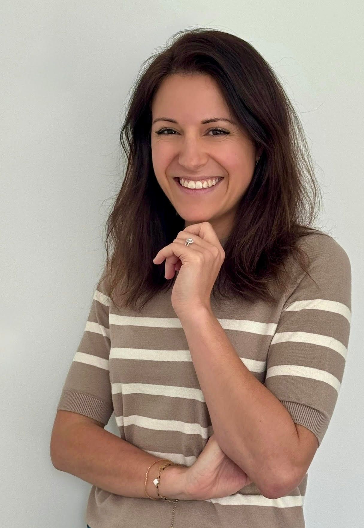 Une femme en chemise rayée sourit, la main au menton, devant un mur blanc.