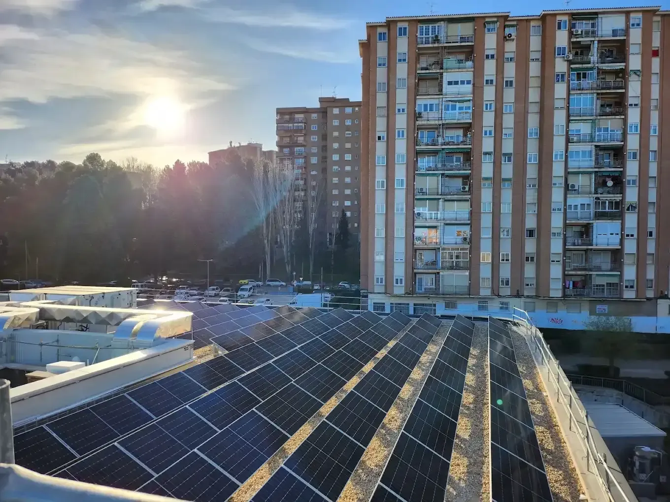 Paneles solares en una azotea al atardecer, con edificios de apartamentos y árboles al fondo.